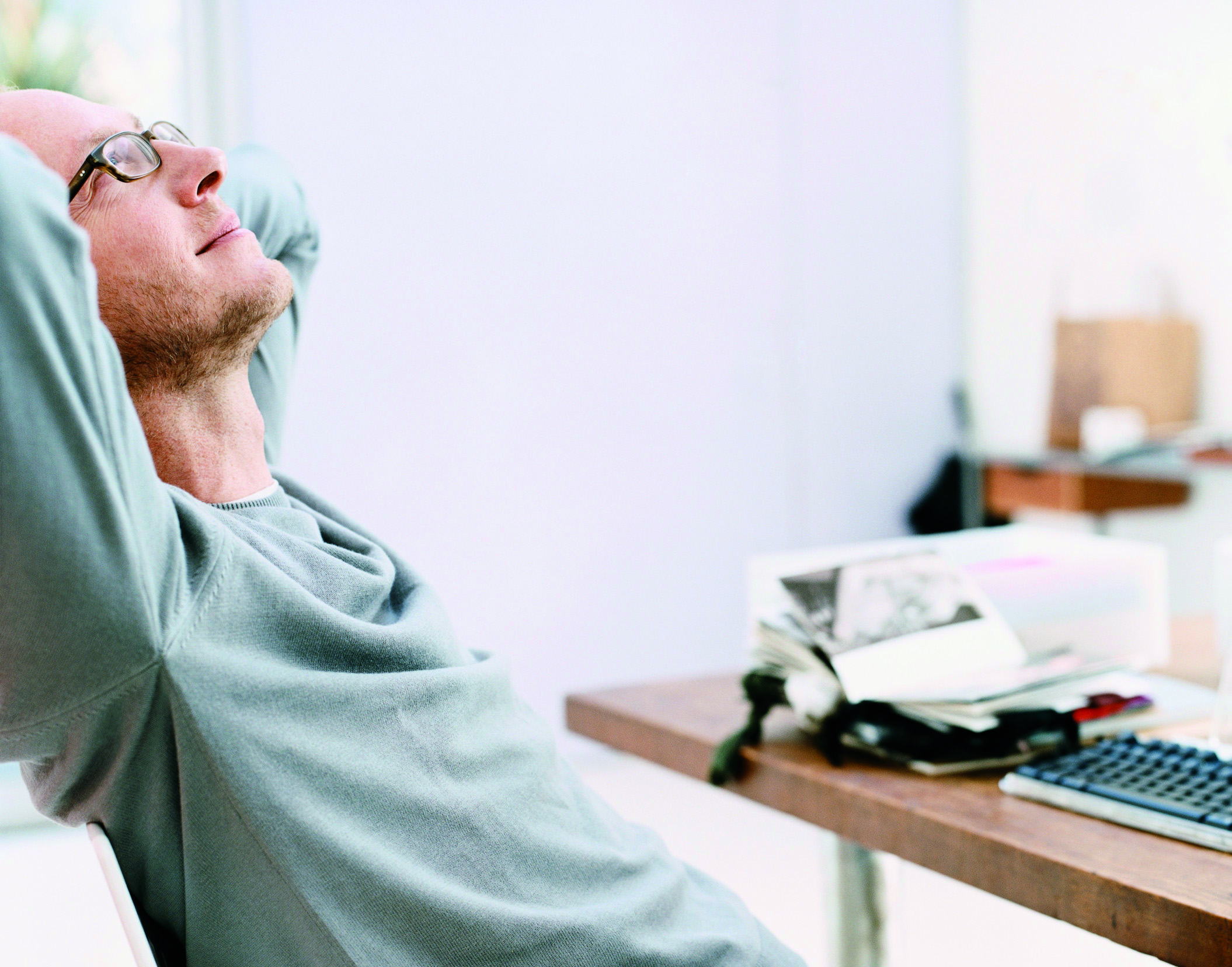 Man Sits at a Desk With His Hands Behind His Head, Looking Up and Daydreaming